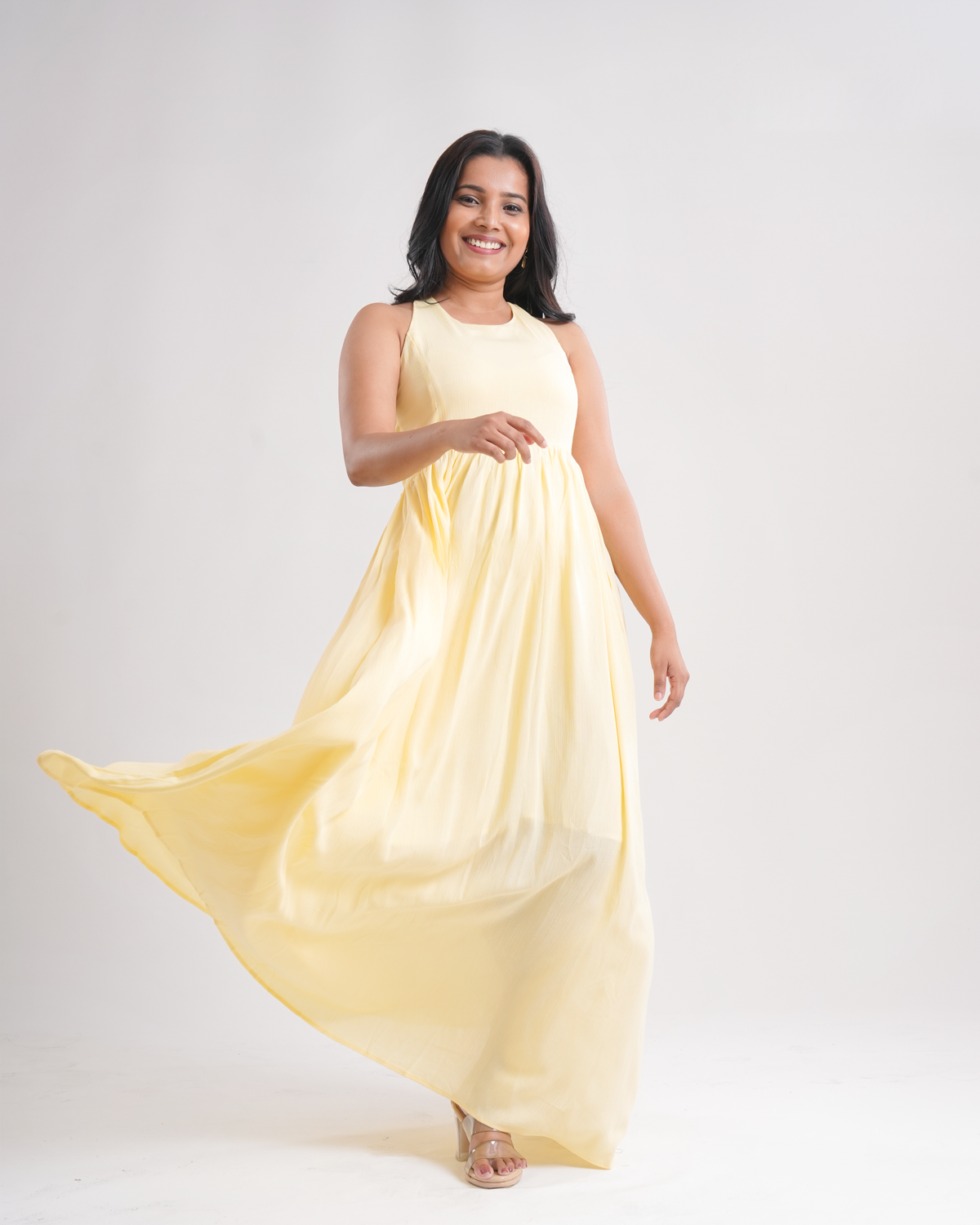 A woman modeling a sleeveless, high-neck, floor-length yellow maxi dress that flows dramatically as she twirls, set against a plain light gray studio background.